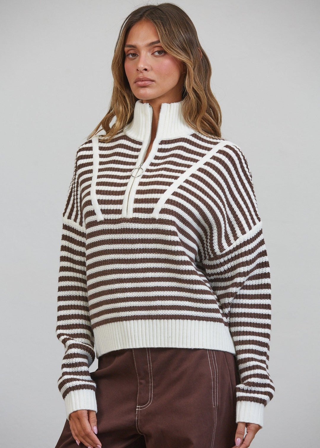 A woman wearing a brown and off-white striped knit mock-neck sweater with a half-zip collar, paired with brown pants, against a solid light gray studio background.