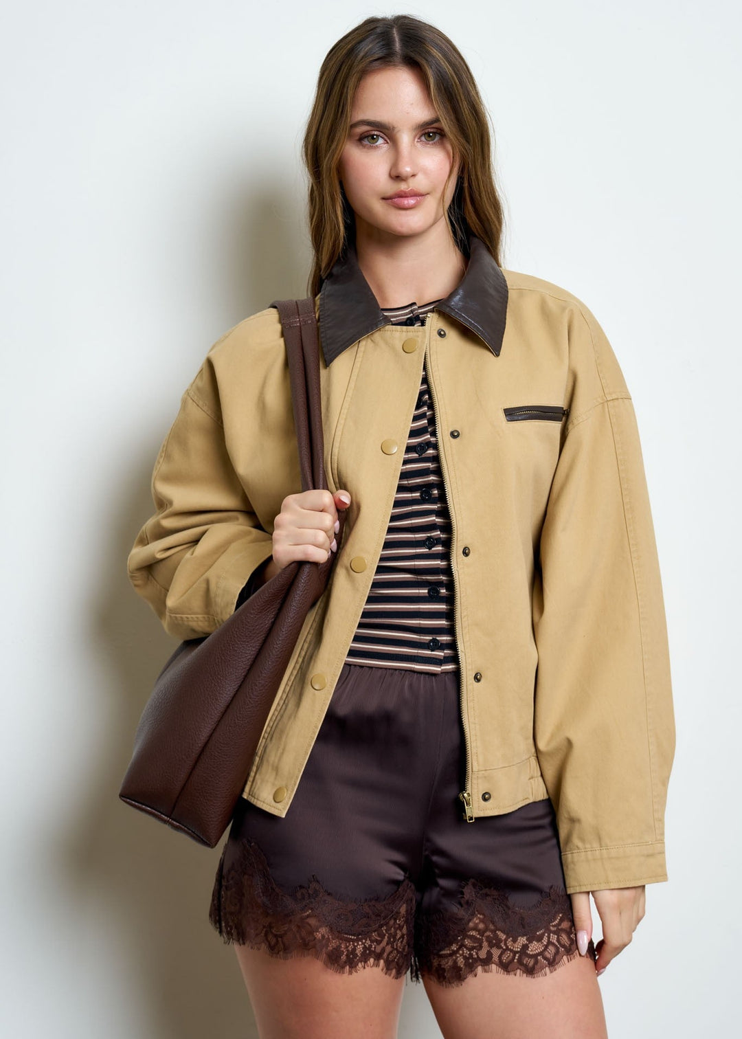 A woman models a tan utility jacket with a dark brown collar, layered over a black and white striped shirt and dark brown lace-trimmed shorts, while carrying a brown leather shoulder bag against a plain light gray background.