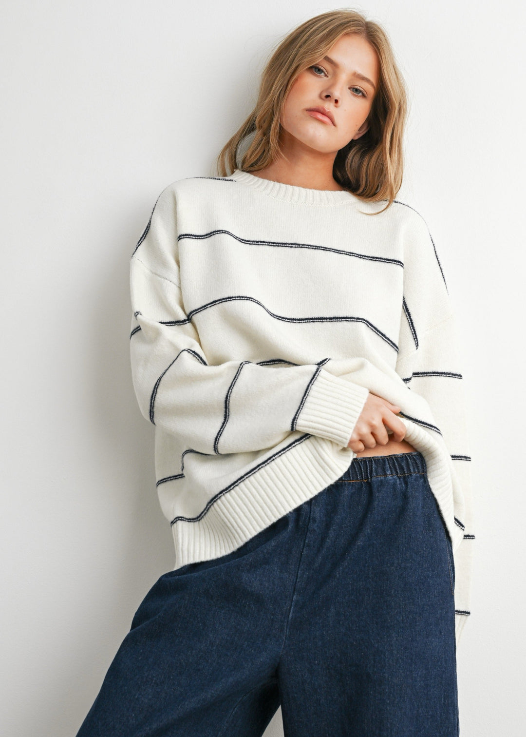 A woman wearing a cream-colored knit sweater with thin black horizontal stripes and dark blue wide-leg jeans, posing against a light gray wall.