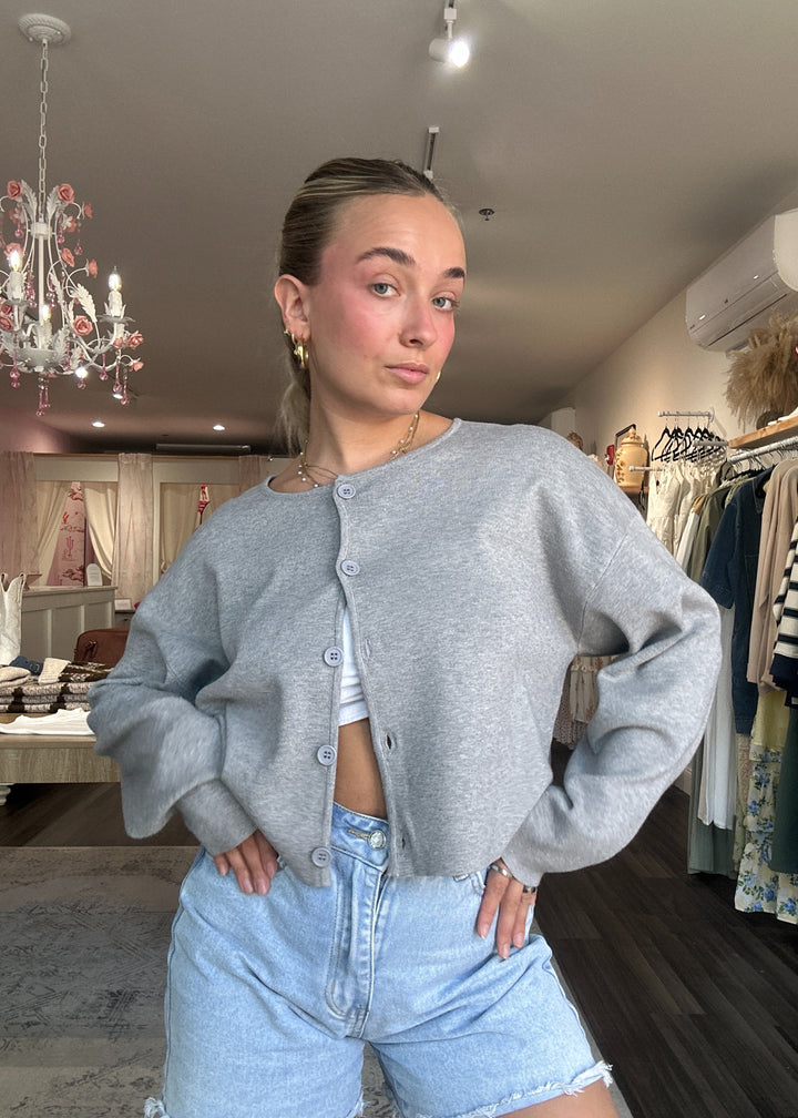 Woman wearing a gray cardigan and light blue jeans in a room with a chandelier and clothing rack.