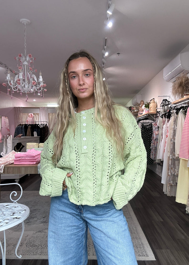 Person wearing a light green sweater and blue jeans standing in a store with racks of clothes.