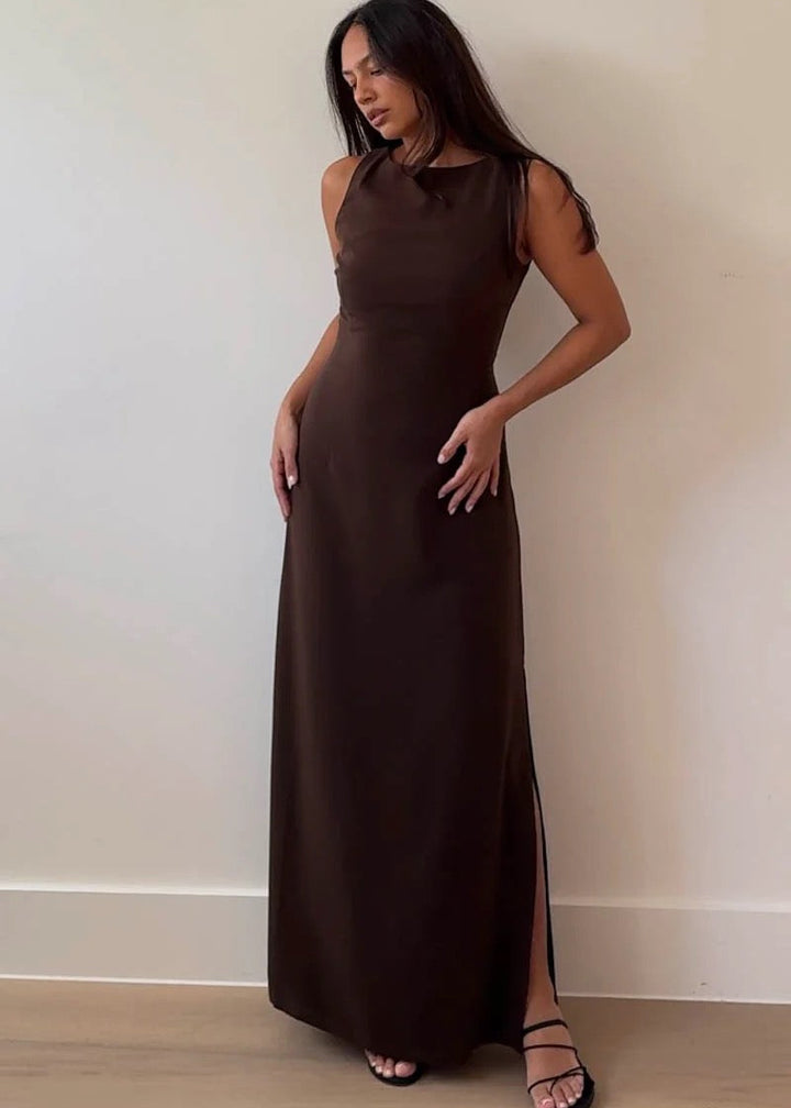 Woman wearing a brown sleeveless dress with a slit, standing against a plain wall.