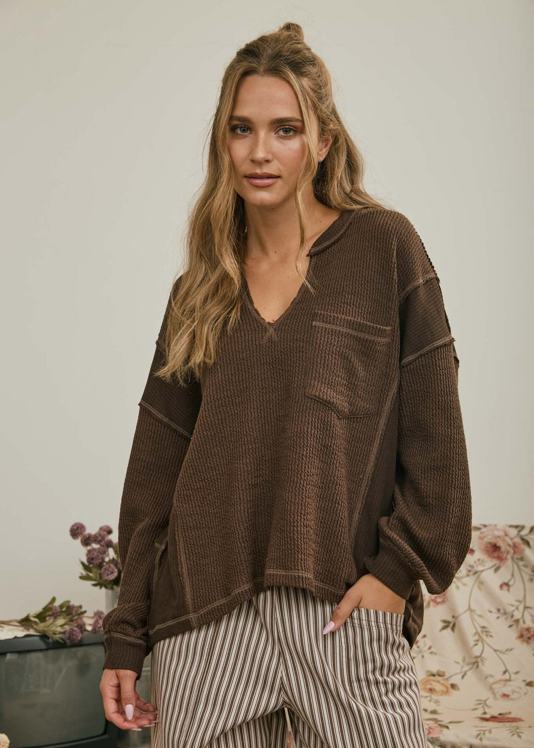 A woman wearing a brown ribbed knit long-sleeve top with a V-neck and striped pants, standing in a softly lit indoor setting.