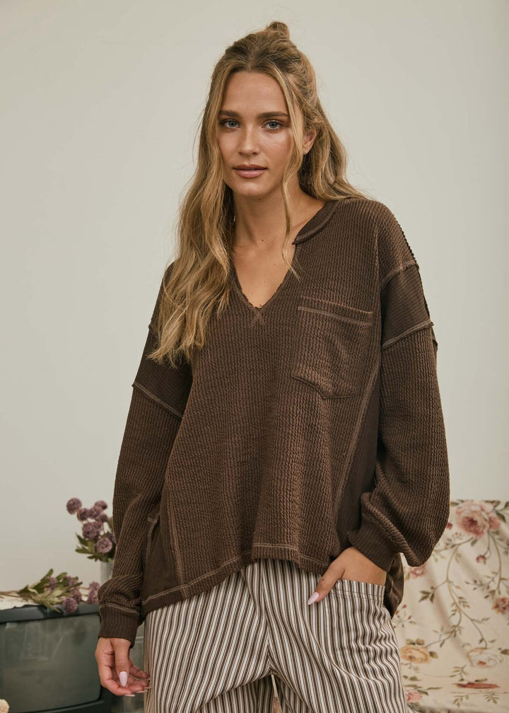 A woman wearing a brown ribbed knit long-sleeve top with a V-neck and striped pants, standing in a softly lit indoor setting.