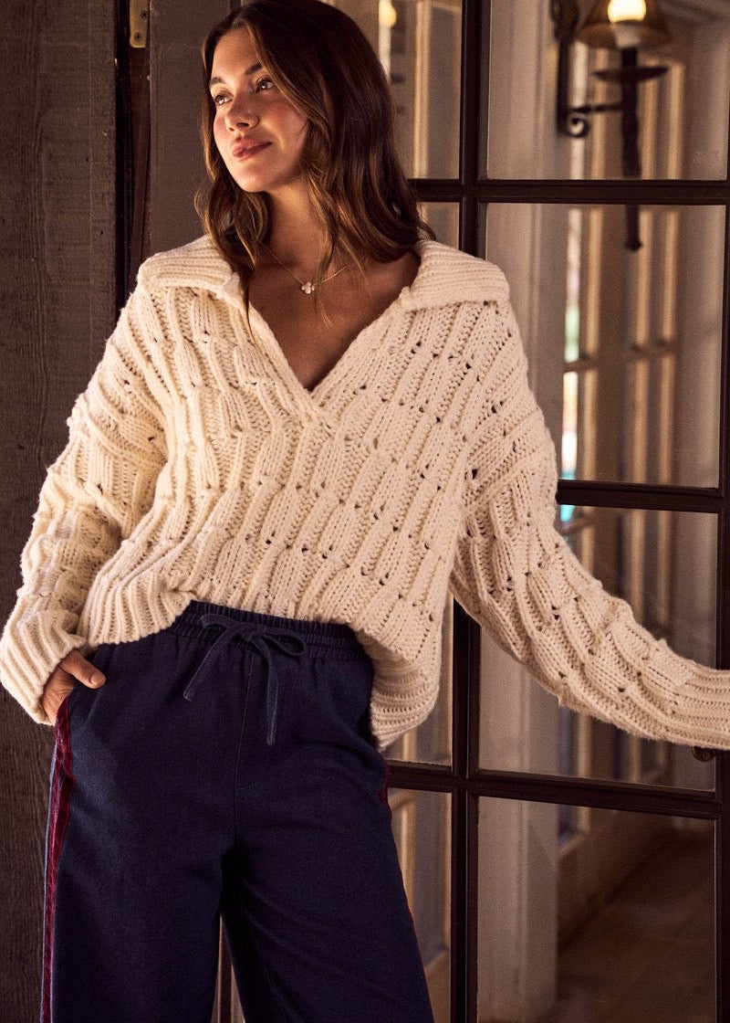 Woman wearing a cream knitted sweater and dark pants indoors.