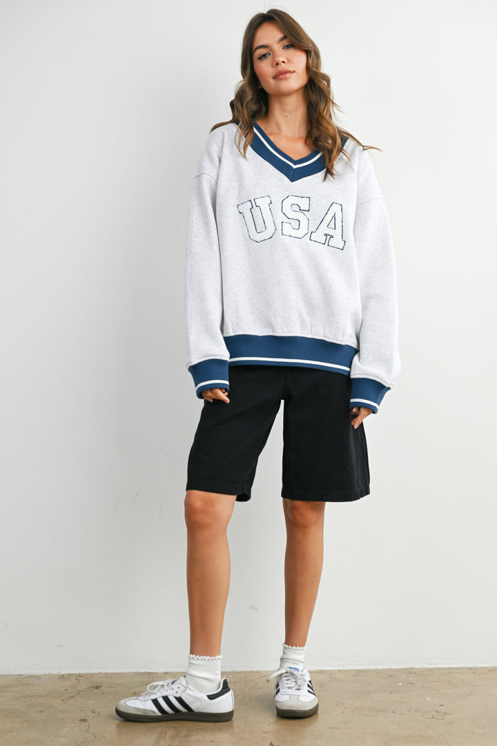 Woman wearing a white 'USA' sweatshirt with blue accents and black shorts on a plain background