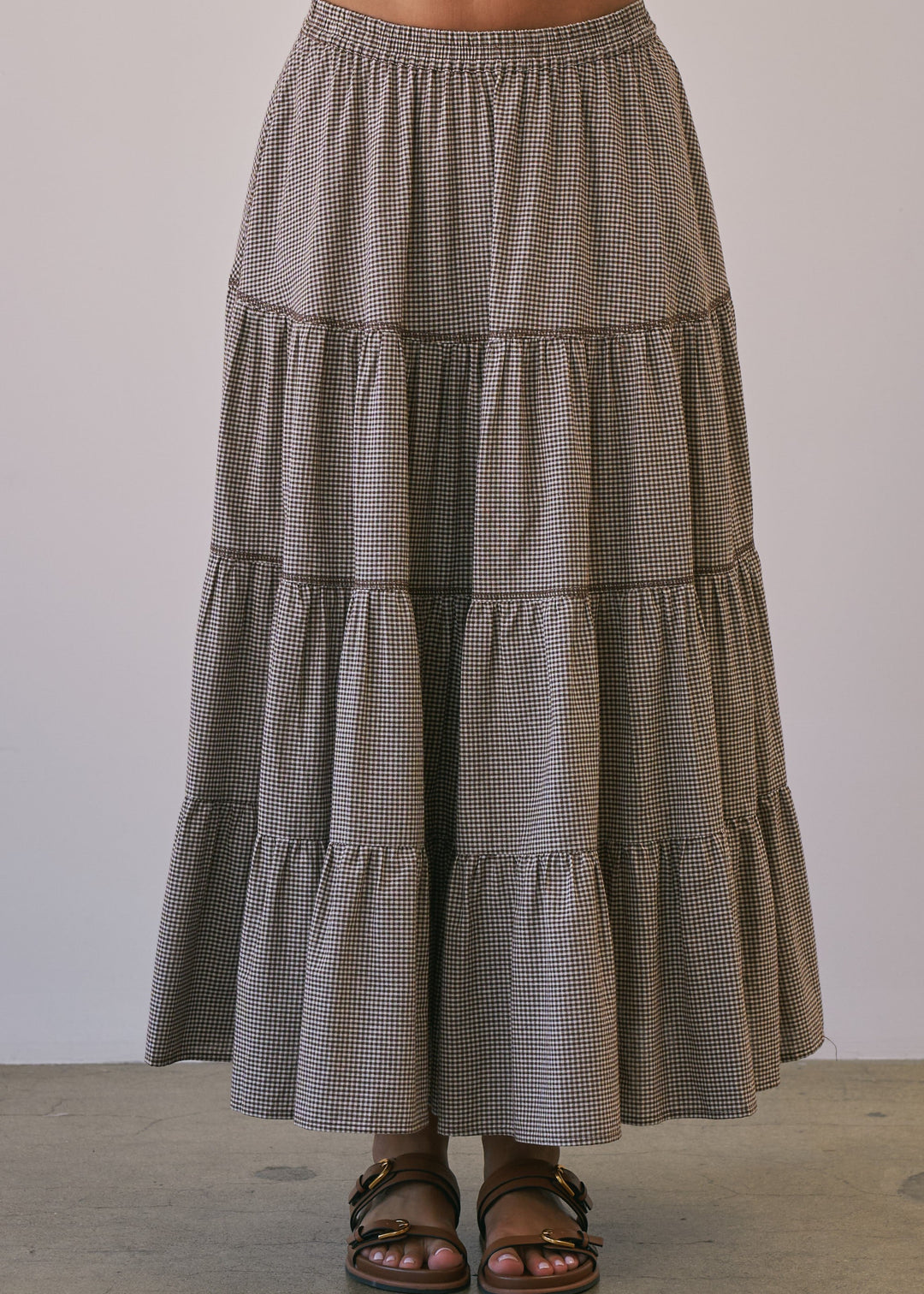 A tiered maxi skirt in a small brown and beige gingham pattern, worn by a model with brown leather slide sandals against a neutral studio background.