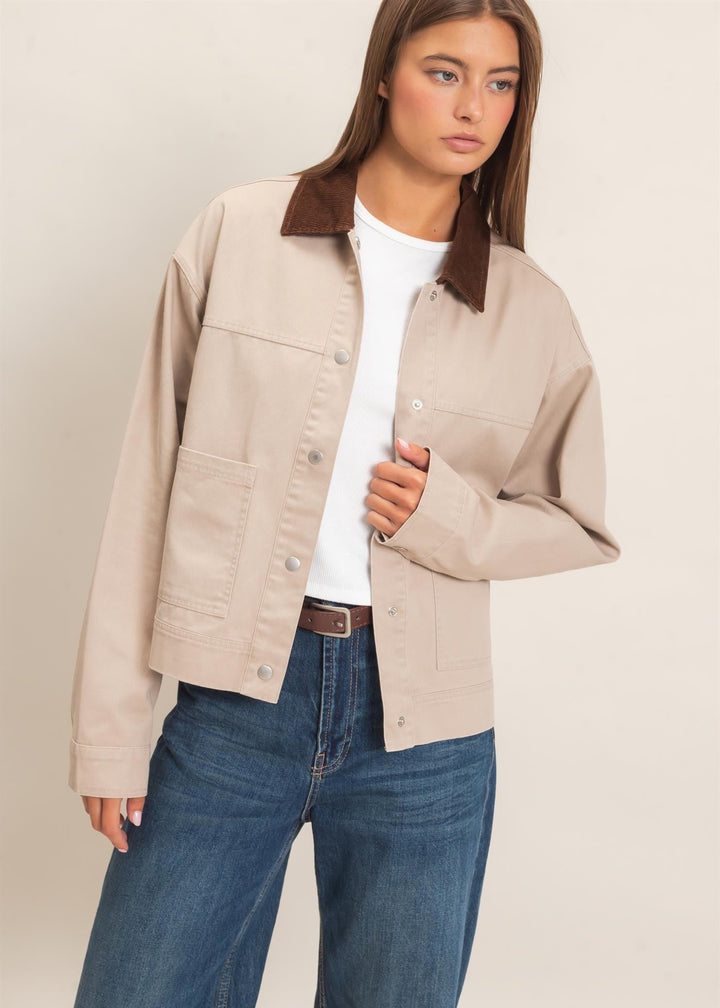 A woman modeling a light beige utility jacket with a contrasting dark brown corduroy collar, worn over a white t-shirt and blue jeans against a plain beige background.